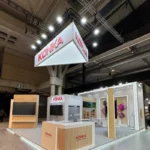 The Konka booth at ISE 2025 in Barcelona, showcasing a modern and sophisticated design with a combination of wood and white materials. The booth is illuminated by spotlights, creating an inviting atmosphere, and features multiple display areas with large screens demonstrating Konka's advanced technological products. A central reception desk is positioned prominently for welcoming visitors, while the brand's overhead signage with the Konka logo underscores its strong presence at the event. The sleek layout reflects Konka's commitment to innovation and high-quality solutions, drawing attention in the bustling trade show environment.
