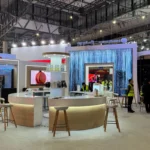 The Liantronics booth at ISE 2025 in Barcelona, showcasing a sleek and innovative design. At the center, a circular counter surrounded by stools provides a welcoming space for visitors. The booth features multiple displays and cutting-edge LED screens, including a captivating decorative waterfall-like LED installation that grabs attention. The area is well-lit, creating a professional and engaging atmosphere. Several team members and attendees are present, reflecting the vibrant activity at the booth. The setup highlights Liantronics' commitment to innovation and advanced display technologies.