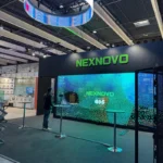 The Nexnovo booth at ISE 2025 in Barcelona, showcasing a futuristic and innovative design. A large, vibrant LED display dominates the booth, featuring the Nexnovo logo prominently displayed in bright green text. Above the setup, a circular LED screen suspended from the ceiling broadcasts dynamic, eye-catching content, capturing the attention of attendees. The booth is situated in a spacious exhibition hall, surrounded by other exhibitors, but stands out due to its advanced technology and interactive setup. A person is seen engaging with the display, emphasizing the hands-on experience that Nexnovo provides. The overall atmosphere highlights Nexnovo's leadership in cutting-edge transparent LED displays and visual solutions.