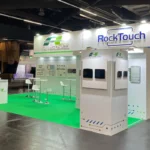 RockTouch booth at Embedded World 2025 in Nuremberg, featuring a clean and modern design with a bright green floor and white walls. The booth prominently displays the RockTouch logo alongside the FutureLabs branding, reflecting a partnership in innovative technologies. Several screens showcase touch and display solutions, highlighting the company's commitment to cutting-edge advancements in interactive technologies. High tables and white chairs create a welcoming space for visitors to engage in discussions, while the overall organization and bright lighting emphasize professionalism and innovation.