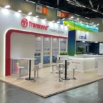 The Transcend booth at Embedded World 2025 in Nuremberg, designed with a professional and welcoming layout, features a white and red color scheme. The prominent Transcend logo at the top is accompanied by the phrase "EMBEDDED SOLUTIONS," highlighting the focus on advanced memory and storage technologies. High tables with white chairs invite visitors for discussions, while the back wall showcases detailed information about cutting-edge solutions like Power Loss Protection, RLC Mode, and TCG Opal. A vertical banner on the left lists product offerings, including SSD Solutions, DRAM Modules, Memory Cards, and Flash Solutions. The well-lit booth, situated in a spacious exhibition hall, draws attention to Transcend's expertise in embedded solutions amidst a vibrant industry environment.
